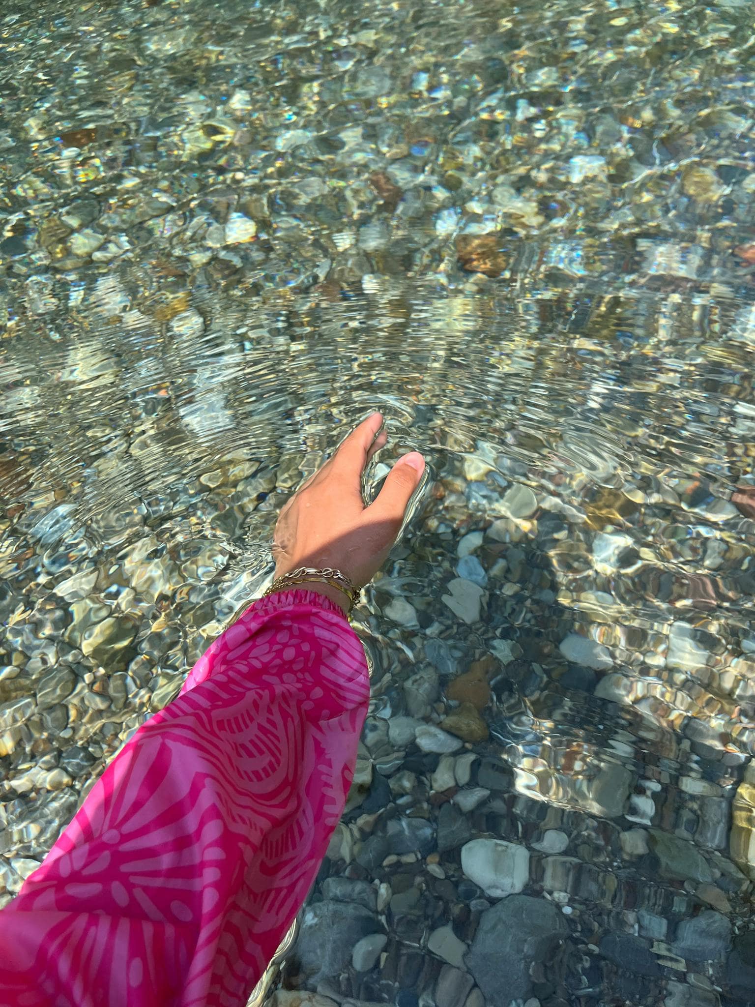 A close-up of a hand in a pink sleeve touching clear, rippling water over river pebbles. Sunlight reflection.
