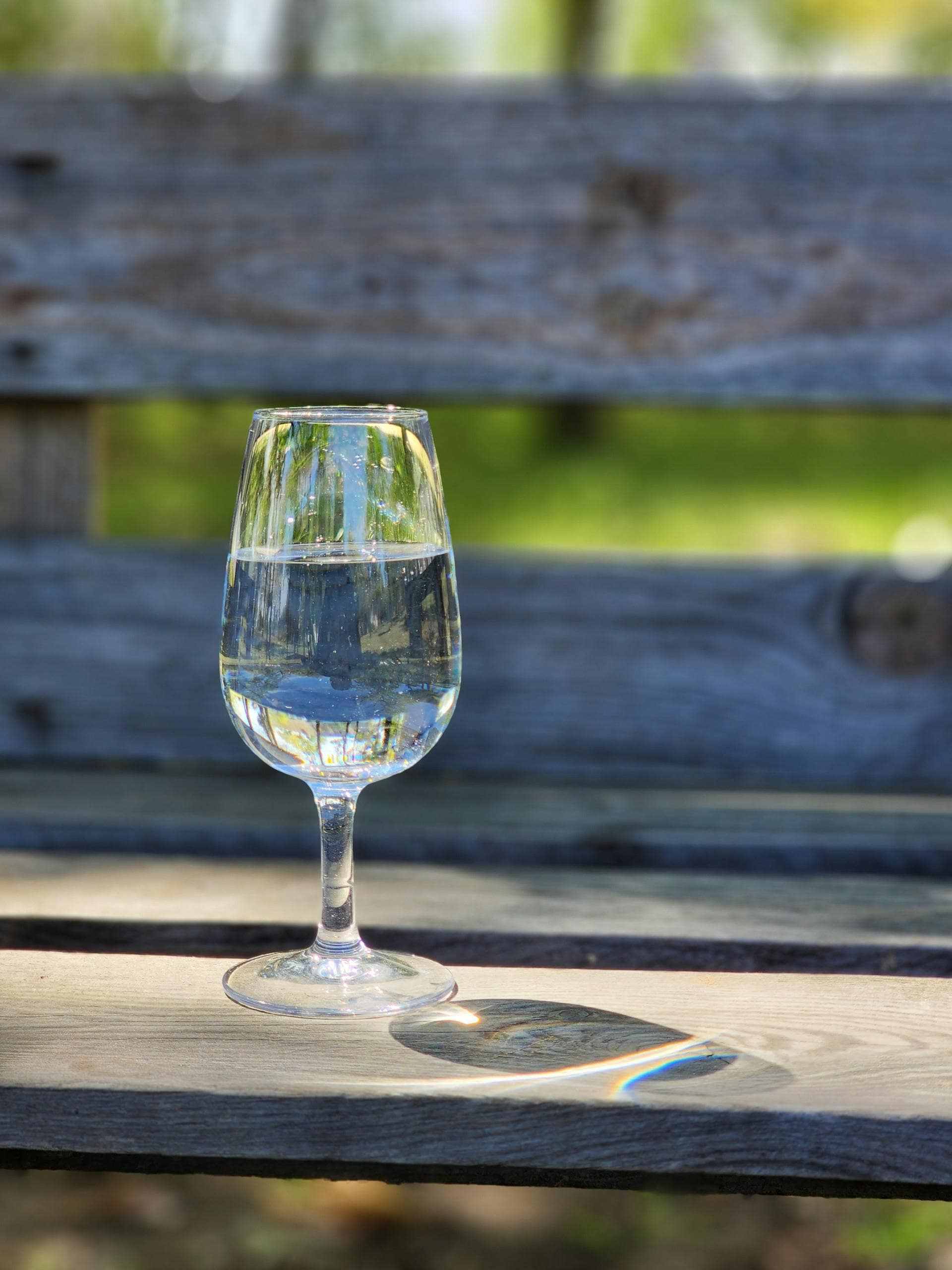 A clear glass of water sits on a wooden bench outdoors, illuminated by sunlight.