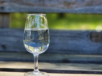 A clear glass of water sits on a wooden bench outdoors, illuminated by sunlight.