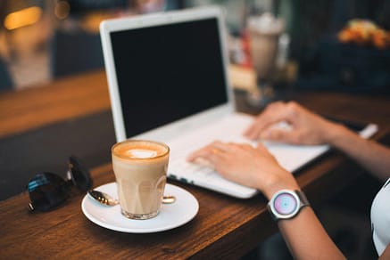 A woman typing on a laptop with a latte on a wooden table, indoors.